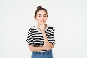 Portrait of cute girl keeps a secret, shushes, shhh gesture, presses finger to lips, gossiping, stands over white background photo