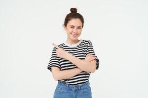 Portrait of smiling, beautiful young brunette woman, pointing finger left, showing advertisement, white studio background photo