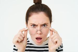 Close up of shocked, insulted young woman, frowning and looking offended, reading smth offensive and upsetting, takes off glasses, stands over white background photo