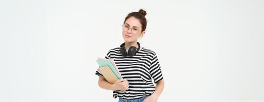 Education concept. Smiling brunette girl, student in casual clothes, holds her books, study material, wears headphones over neck, looks confident and happy, isolated over white background photo