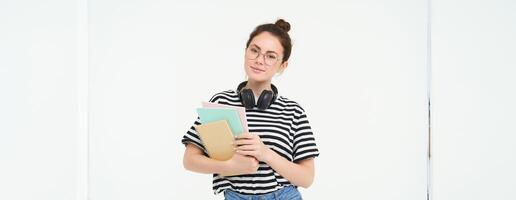 Student and education concept. Young woman with books, notes and pen standing over white background, college girl with headphones over neck posing in studio photo