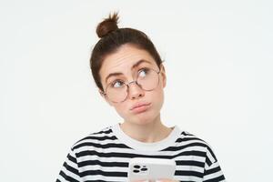 Close up portrait of girl in glasses, looks puzzled, holds phone and thinks, makes choice, decides what to order, does online shopping in mobile app, white background photo