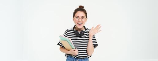 Portrait of excited young woman in glasses, looks surprised and happy. Student standing on white background with books and notebooks photo