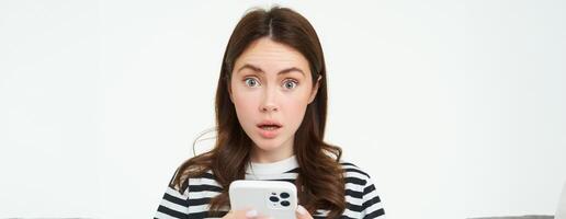 Portrait of woman with smartphone in hands, looking surprised, raising eyebrows from amazement, white background photo