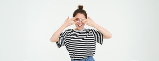 Happy young woman, bday girl peeking through fingers, holds hands on eyes, waiting for surprise, stands over white background photo