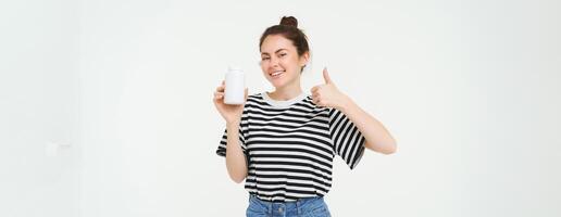 Portrait of beautiful, smiling brunette woman, shows thumbs up and bottle of vitamins, recommends dietary supplements, isolated against white background photo