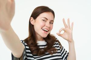 retrato de positivo, contento joven mujer muestra bueno firmar y toma autofoto, sostiene teléfono inteligente con extendido mano, posando cerca alguna cosa ella recomienda, aislado en blanco antecedentes foto