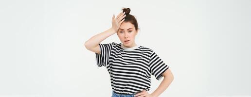 Shocked woman, slaps her forehead, holds hand on head and looks startled, concerned re something bad, terrible situation, standing over white background photo