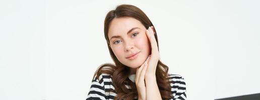 Portrait of tender, beautiful young woman, touches her clear, glowing skin, without blemishes or acne, concept of makeup and cosmetology, white background photo