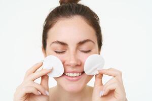 Image of young woman taking off her makeup with cotton pads, using facial cleanser, cleaning her face with skincare treatment, standing over white background photo