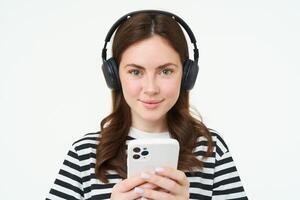 imagen de morena joven mujer, sonriente, escuchando a música en auriculares, acecho videos en móvil teléfono aplicación, participación teléfono inteligente, en pie terminado blanco antecedentes foto