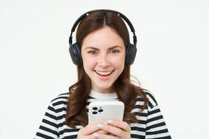 imagen de joven mujer en auriculares, utilizando teléfono inteligente y reír, acecho vídeo en móvil teléfono, escucha a música en transmisión Servicio aplicación, blanco antecedentes foto