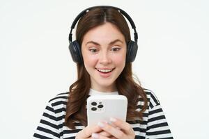 Young woman with surprised face, looking at smartphone screen, reacts amazed, looks with happiness at mobile phone, standing in wireless headphones, white background photo