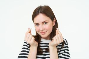 retrato de linda joven morena mujer, muestra kawaii dedo corazones y sonriente, gustos algo, soportes en a rayas camiseta terminado blanco antecedentes foto