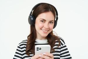 imagen de morena joven mujer, sonriente, escuchando a música en auriculares, acecho videos en móvil teléfono aplicación, participación teléfono inteligente, en pie terminado blanco antecedentes foto