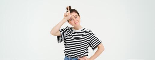 Portrait of pleased, arrogant young woman, showing loser gesture, l letter on forehead, smirking, standing over white background photo