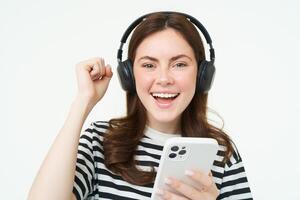 entusiasta niña en auriculares, participación teléfono inteligente, sonriente y aplausos, celebrando, levantamiento mano arriba, lograr meta, en pie terminado blanco antecedentes foto