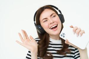 tecnología y social medios de comunicación. retrato de contento joven mujer canto karaoke, jugando música aplicación en teléfono inteligente, vistiendo inalámbrico auriculares, en pie terminado blanco antecedentes foto