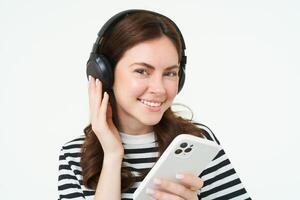 retrato de contento joven moderno mujer, hembra modelo en inalámbrico auriculares, sonriente y mirando a cámara, participación teléfono inteligente, utilizando móvil teléfono aplicación, blanco antecedentes foto