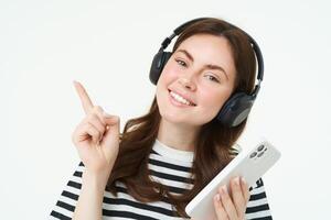 retrato de sonriente alumno, niña en auriculares, participación móvil teléfono, señalando izquierda, demostración anuncio publicitario, Tienda oferta, blanco antecedentes foto