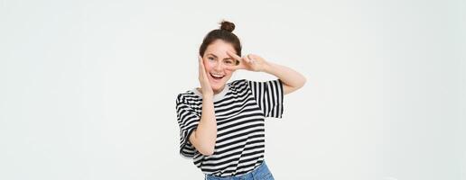 linda europeo mujer posando para foto, participación paz, kawaii mano gesto cerca rostro, sonriente a cámara, en pie terminado blanco antecedentes foto