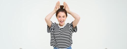 Enthusiastic young woman, holding hands on top of head, animal ears gesture, laughing and smiling, isolated against white background photo