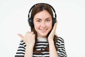 Music store concept. Smiling young woman listening song in headphones, pointing left at copy space, shows advertisement text, stands over white background photo