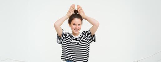 Image of beautiful, happy young woman showing bunny ears on top of her head, looking excited, posing over white background photo