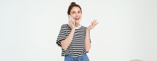 Happy young woman talks on mobile phone, chats on telephone, uses smartphone, stands over white background photo