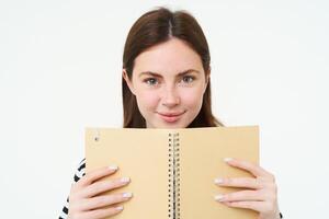 Image of young woman with notebook, holding her personal diary, work planner, smiling, reading something, standing over white background photo