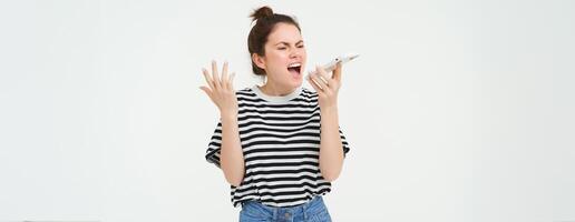 Annoyed, angry young woman shouts at speakerphone, yelling at mobile phone and shaking hands, standing over white background photo