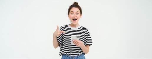 Portrait of beautiful young woman, recommending app on the phone, pointing at mobile screen, showing smth on smartphone, standing against white background photo