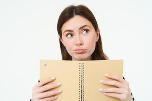 Portrait of thinking girl holding a notebook. Young woman with thoughtful face, holding weekly planner, checks her schedule, writes notes in her memo book, stands over white background photo