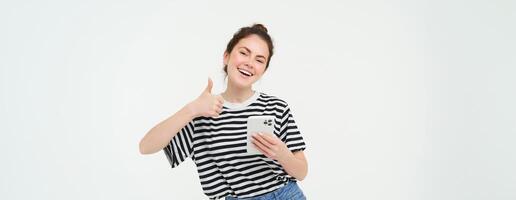 entusiasta niña con teléfono inteligente muestra pulgares arriba, aislado terminado blanco antecedentes foto