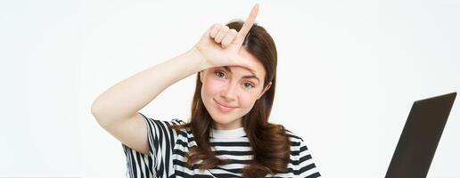 Close up portrait of young brunette girl mocking you, showing loser finger gesture, l letter with hand on forehead, smiling with pleased face, standing isolated on white background photo