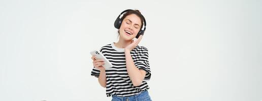 Young woman with smartphone listening to music, dancing to her favourite song in headphones, posing against white background photo