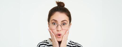 Image of surprised cute girl in glasses, holds hands on face, says wow, impressed by smth, isolated over white background photo