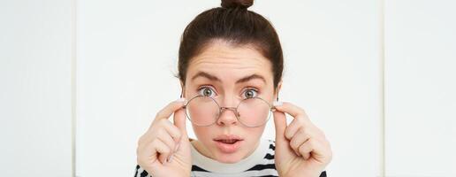 retrato de mujer propensión hacia cámara como Si leyendo texto con sorprendido rostro, toma apagado anteojos, soportes terminado blanco antecedentes foto