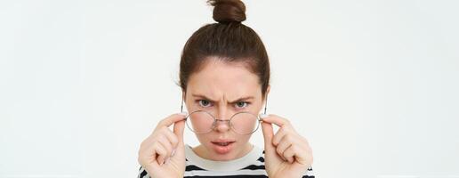 Portrait of shocked, angry woman frowning, looks confused at camera, takes off glasses with frustrated face expression, stands over white background photo