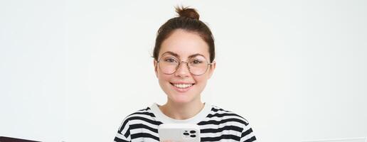 retrato de hermosa joven mujer en anteojos, participación teléfono inteligente, utilizando móvil teléfono, en línea compras en solicitud, en pie terminado blanco antecedentes foto