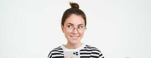 imagen de joven mujer en anteojos, utilizando su móvil teléfono, en pie con teléfono inteligente y sonriente, en pie terminado blanco antecedentes foto