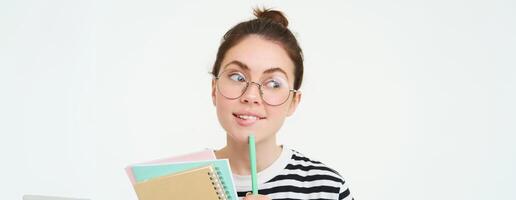 Portrait of cute woman, student in glasses, thinking, standing thoughtful with notebooks, memo notes, white background photo