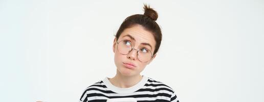 Close up portrait of girl in glasses, looks puzzled, holds phone and thinks, makes choice, decides what to order, does online shopping in mobile app, white background photo