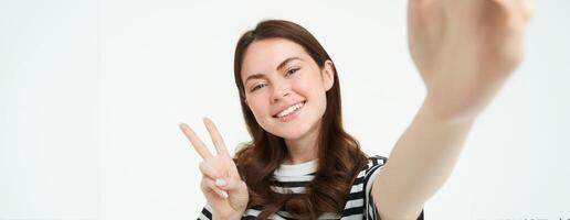 Smartphone view, woman holding mobile phone camera and taking selfie with peace, v-sign gesture, smiling at camera, posing over white background photo