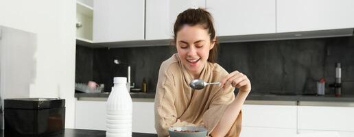 Portrait of happy young woman leans on kitchen worktop and eating cereals, has milk and bowl in front of her, having her breakfast, wearing bathrobe photo