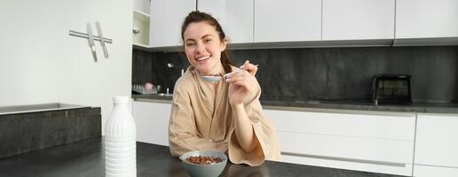 Portrait of happy young woman leans on kitchen worktop and eating cereals, has milk and bowl in front of her, having her breakfast, wearing bathrobe photo