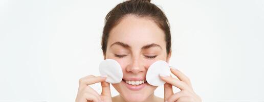 Image of young woman taking off her makeup with cotton pads, using facial cleanser, cleaning her face with skincare treatment, standing over white background photo