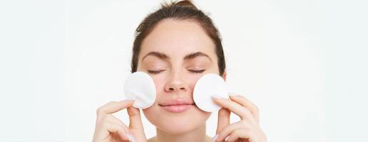 Image of young woman taking off her makeup with cotton pads, using facial cleanser, cleaning her face with skincare treatment, standing over white background photo