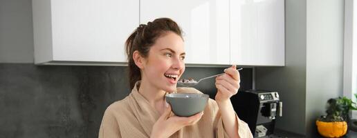 Gorgeous girl holds spoon near mouth, eats cereals with milk, leans on kitchen worktop photo