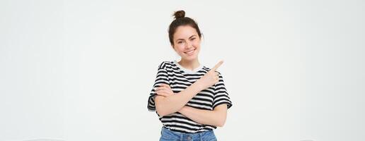 retrato de linda sonriente hembra modelo, vistiendo casual ropa, señalando dedo izquierda, demostración anuncio publicitario, blanco antecedentes foto